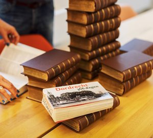 Op  tafel in de studiezaal ligt een stapel boeken, waaronder Dordrecht in 365 dagen. Twee handen bladeren door een boek.