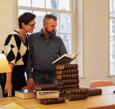 Een man en vrouw staan de de studiezaal van het Regionaal Archief Dordrecht. Ze bladeren in een boek. Voor hen ligt een stapel boeken op tafel.