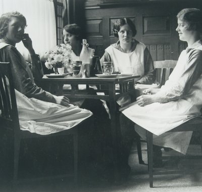 Vier dames in werkschorten zitten aan een rond tafeltje. Er staan kopjes en schoteltjes en een kan op tafel.