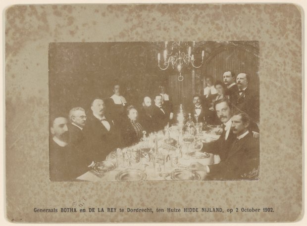 Grote groep mannen en enkele vrouwen aan een rijk gedekte tafel.