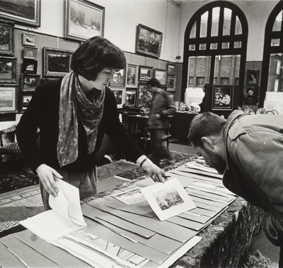 In een ruimte met vele schilderijen aan de muur staan een man en vrouw gebogen voor een tafel terwijl ze foto's bestuderen.