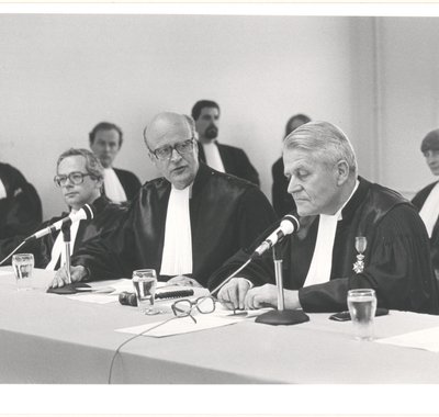 Drie rechters aan een bureau met microfoons. Achter hen zitten nog 5 mensen, allen in toga.