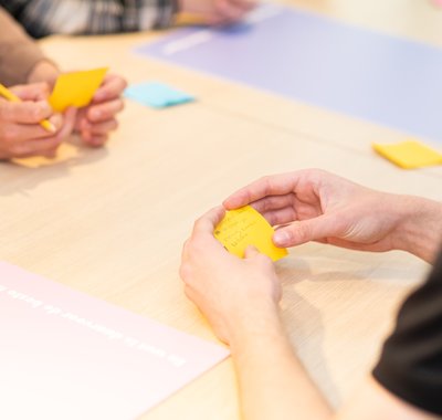 Kinderen zitten aan tafel, je ziet alleen hun handen. Op Post-its hebben ze wat geschreven.