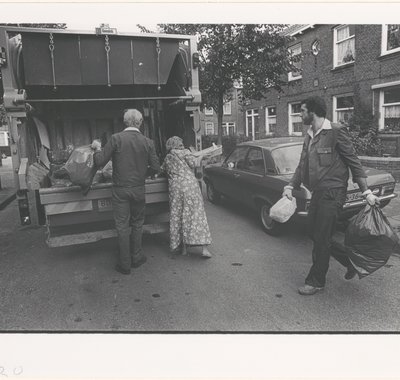 De vuilniswagen komt langs. Twee mannen en een vrouw gooien hun vuilnis erin.