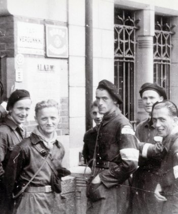 Negen mannen, lid van de Binnenlandse Strijdkrachten, staan lachend op de foto voor een bankgebouw.