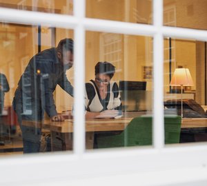 Door het buitenraam kijken we de studiezaal Regionaal Archief Dordrecht in. Binnen bestuderen een man en vrouw documenten.