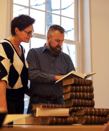 Een man en vrouw staan de de studiezaal van het Regionaal Archief Dordrecht. Ze bladeren in een boek. Voor hen ligt een stapel boeken op tafel.
