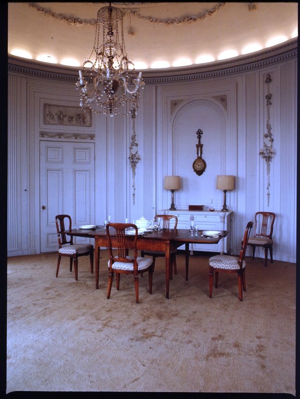 Rijkelijk gedecoreerde kamer met een tafel en stoelen en theepot. Er hangt een kroonluchter.